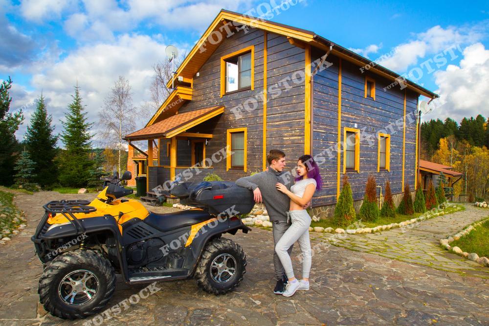 На фотографии мужчина и женщина стоят рядом с квадроциклом возле деревянного загородного дома. Пара улыбается и общается, наслаждаясь отдыхом в красивой природной обстановке. Снимок символизирует активный отдых, романтику и свободу на природе.